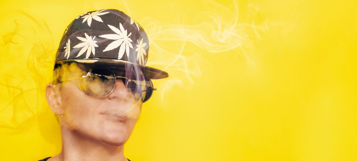 Girl in sunglasses and cap with leaves of marijuana smokes on yellow background